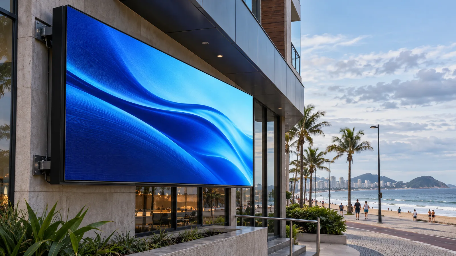 Painel de LED em fachada comercial próxima ao mar, com praia e calçadão ao fundo.